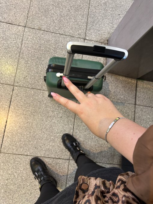The image shows a person's feet wearing black boots, holding a green suitcase with wheels and a handle while making a peace sign with their fingers. The caption of the image reads "OMW to Solana Summit Berlin @SuperteamDE".