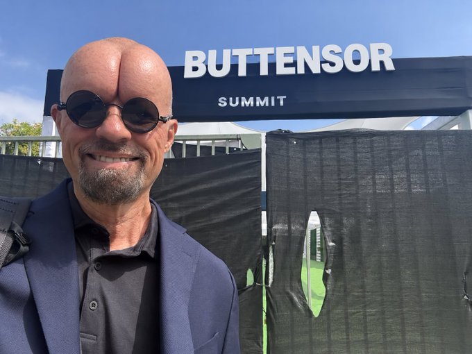 A selfie of a bald man with round sunglasses and a beard smiling in front of a 'BUTTENSOR SUMMIT' sign.