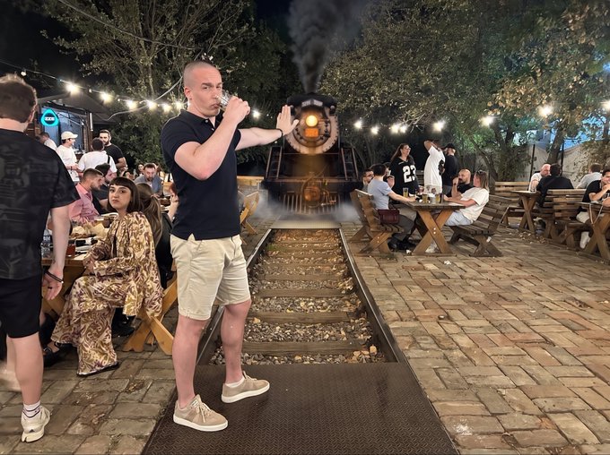 A nighttime outdoor scene at a railway-themed bar or restaurant where a man in a black polo shirt and khaki shorts drinks from a small cup while holding his hand up toward an approaching train with a glowing headlight on tracks surrounded by seated patrons.