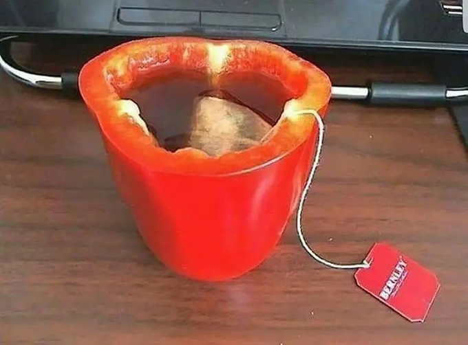 A hollowed-out red bell pepper is used as a cup for steeping a Lipton tea bag, with the string and tag hanging over the side onto a wooden surface.