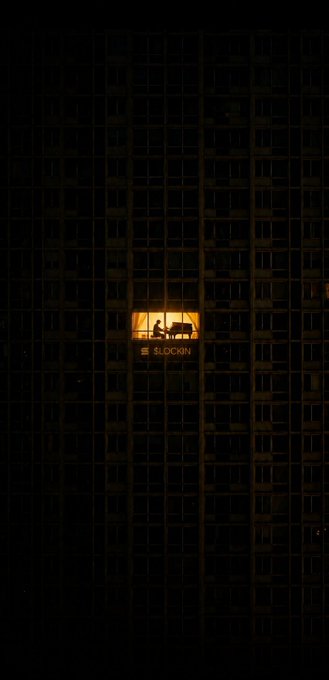 An artistic nighttime photo of a large dark building with a single brightly lit window showing the silhouette of a person and the word 'SLOCKIN' on the window.