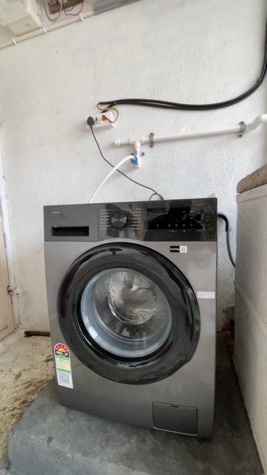 A dark gray front-loading washing machine installed in a basement or utility room with white textured walls, visible PVC plumbing pipes, and electrical wiring above it.