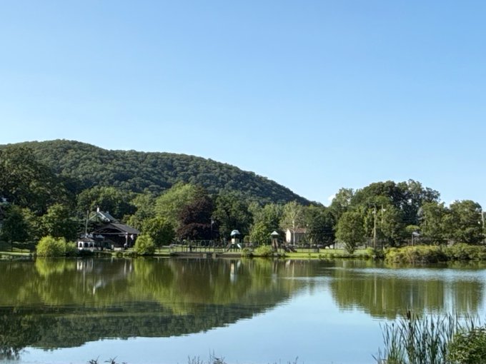A serene landscape photograph showing a calm pond reflecting a clear blue sky, with lush green mountains, trees, and houses lining the far shore.