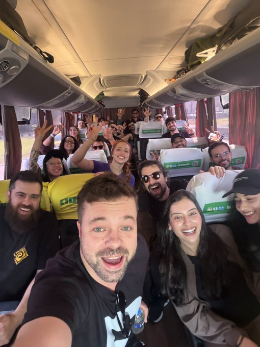 A group selfie taken inside a tour bus with a large group of young adults smiling, cheering, and raising their hands enthusiastically.