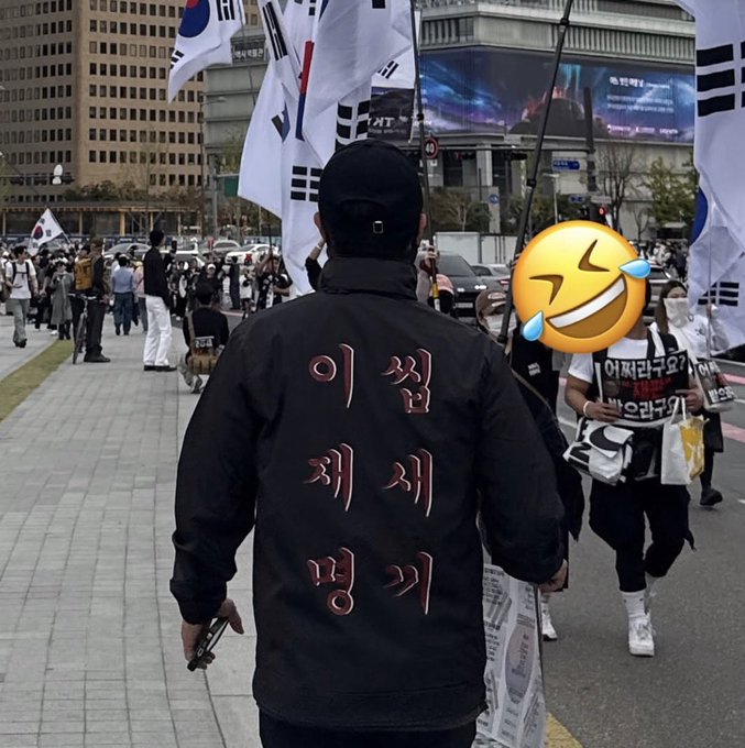A man in a black jacket with Korean text on the back ("이쒸 쳐제 뺑끼") walks through a street protest featuring South Korean flags and demonstrators carrying signs that read "허재라구요" and "안이라구." The post's frustrated comment about not knowing the person's name aligns with the image showing a protester whose identity remains obscured.