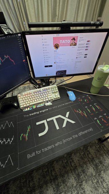 A trading desk setup with dual monitors showing charts and a website, accompanied by a 'JTX' branded desk mat reading 'The trading engine for who you become' and 'Built for traders who know the difference'.