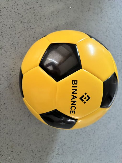 A yellow and black soccer ball featuring the "BINANCE" logo and emblem rests on a gray surface, visually echoing the post's metaphor of "EXchange The World" by transforming a symbol of global sport into a representation of worldwide exchange.