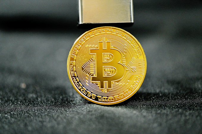 A close-up photograph of a gold Bitcoin coin resting on a dark textured surface, with metallic tongs holding it from above. The coin's inscription includes "BITCOIN DIGITAL DECENTRALIZED PEER TO PEER" and "FINE COPPER."