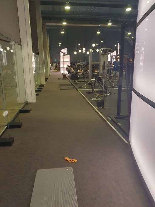 An indoor walkway or corridor alongside a glass-walled gym, showing exercise equipment inside and a yoga mat with an orange strap on the carpet.