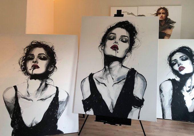 A collection of black-and-white charcoal-style portrait paintings of women with dark eye makeup, displayed on easels in an artist's studio, alongside a printed reference photo in the background.