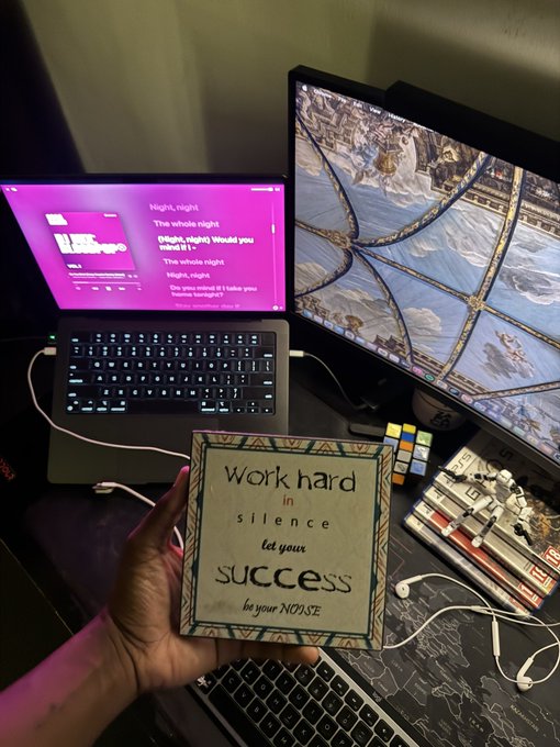A hand holds up a framed motivational sign reading "Work hard in silence, let your success be your NOISE," set against a dual-monitor workspace displaying lyric and a Sistine Chapel ceiling view. The image visually echoes the post's triumphant tone, symbolizing the quiet grind behind the shared $5k fees, $1k distribution, and $2k token burn.
