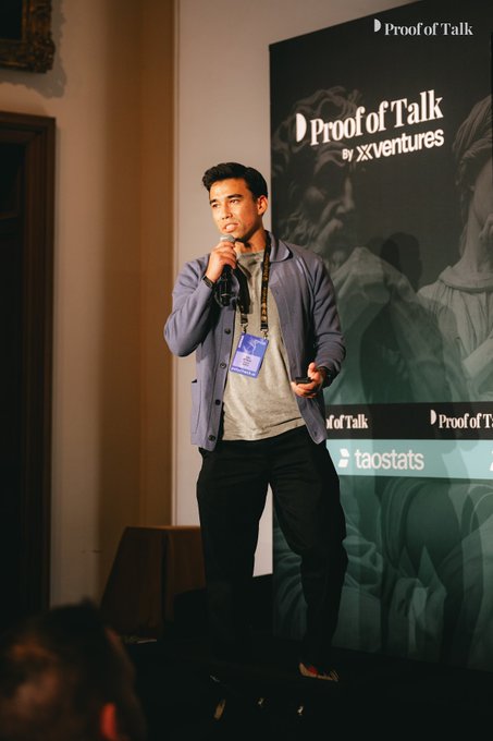 The image shows a man standing on stage at an event, giving a speech or presentation. He appears to be wearing a blue jacket and black pants. The background features a banner with white text that reads "Profit of Talk" and "X-ventures". There are also other people present in the scene, suggesting this is a public gathering or event.