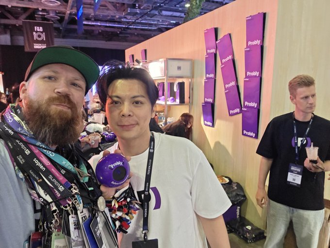 Two attendees pose for a selfie at a conference booth, with the person on the right holding a purple “Probly” magic 8 ball. Probly-branded purple wall signs, lanyards, and event badges are visible, matching the post about using ProblyHQ’s magic 8 ball to get Ron in the photo.