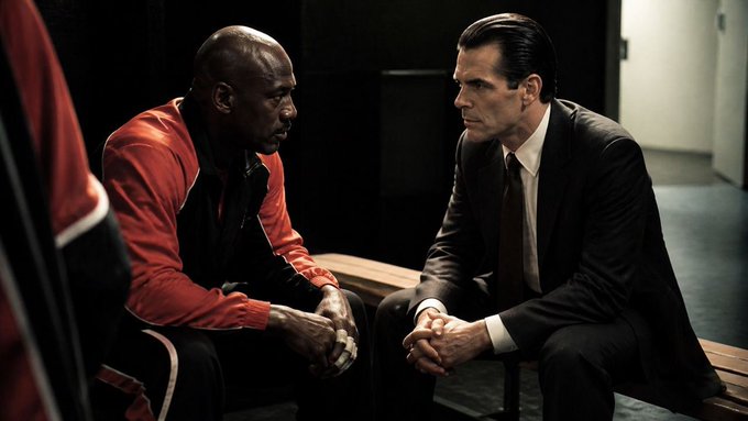 Two men sit on a bench in a dimly lit locker room; one wears a red and black athletic jacket while the other wears a dark suit and tie.