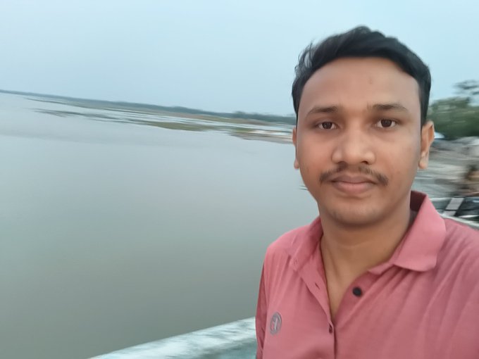 The image features a man wearing a pink shirt standing near a body of water with his hands at his sides. The man appears to be posing for a photo, possibly for a social media post or an event related to Web3 technology. In the background, there are trees and other people present, suggesting that this location might be a popular spot for gatherings or outdoor activities.