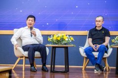 Two men sitting on chairs on a stage during what appears to be a panel discussion or interview, with a small table with yellow flowers between them and a blue digital backdrop.