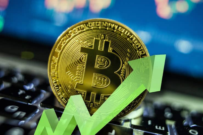 A photograph of a gold Bitcoin token placed on a laptop keyboard with a green upward-pointing arrow graphic overlaid, symbolizing rising cryptocurrency prices and digital asset investment.