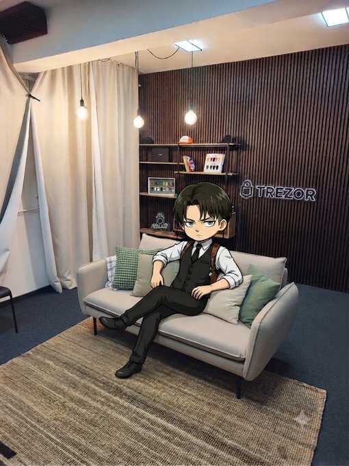A chibi-style illustration of a dark-haired boy wearing a vest, tie, and shoulder holster, sitting on a beige couch in a modern room with a 'TREZOR' sign on the wooden wall.