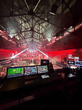 A view from behind a stage control setup with monitors, equipment, and red lighting, looking out toward a stage with a large dome structure overhead.
