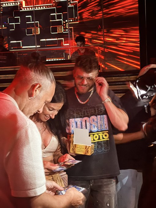 A man in a black graphic t-shirt at what appears to be a nightclub or event venue, smiling while holding what looks like a promotional card, with other attendees gathered around.