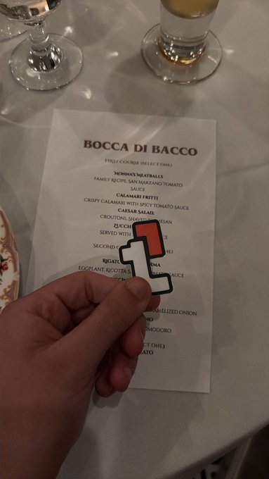A hand holds a small numbered paddle (showing '1') in front of a Bocca di Bacco restaurant menu, with wine glasses visible on the table.