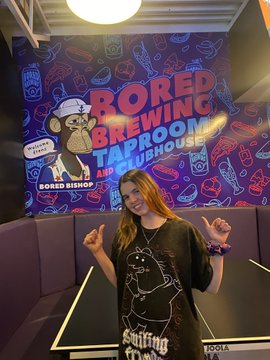 A smiling woman poses in front of a colorful wall mural reading 'BORED BREWING TAPROOM AND CLUBHOUSE' featuring a cartoon character and a 'Bored Bishop' mascot, with her making peace signs.