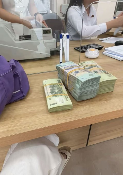Bank teller counter scene showing stacks of Vietnamese dong banknotes (100,000 VND denominations) bound with rubber bands on the desk, with a currency counting machine and bank employees visible in the background.
