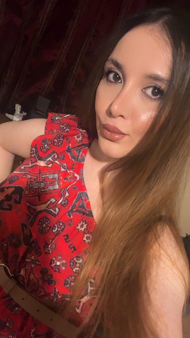 A close-up selfie of a woman with long brown hair wearing a red patterned halter top and a beige belt.