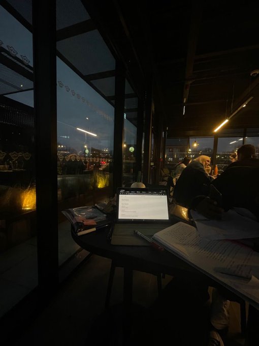 The image shows an open laptop computer sitting on top of a table, with several books scattered around it. The scene takes place at night, with people seated at tables nearby. A person wearing a black jacket can be seen in the background, possibly enjoying their time at the restaurant or bar. The post appears to be promoting a new app called "quip" that allows users to access both classical and quantum computing power for free.