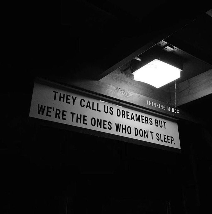 A black and white photo of an illuminated sign hanging from a wooden ceiling that reads 'THEY CALL US DREAMERS BUT WE'RE THE ONES WHO DON'T SLEEP.' with 'THINKING MINDS' written above it.