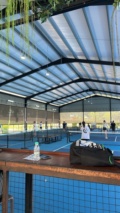An indoor pickleball court with a high metal roof where several people are playing, with a bench in the foreground holding a water bottle, a phone, and a duffel bag.
