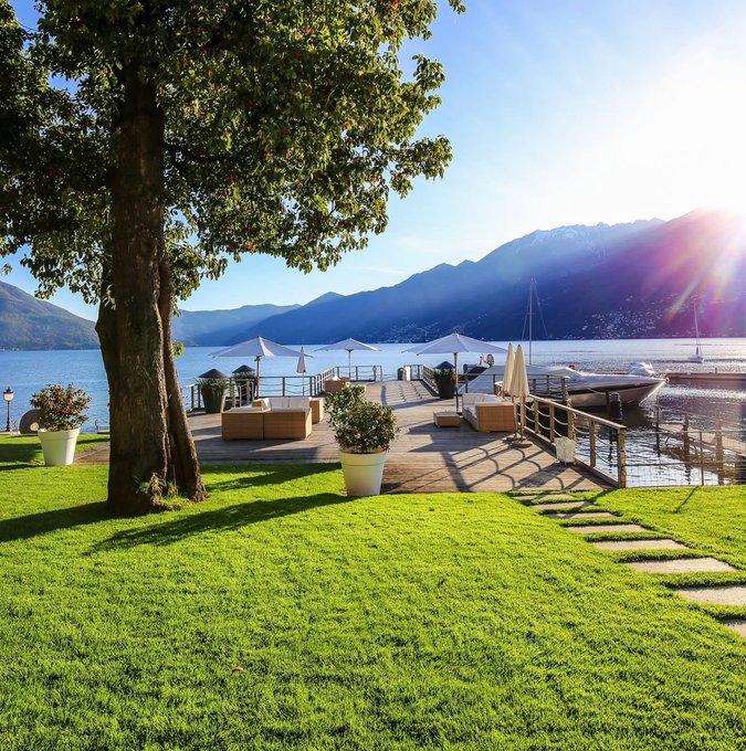 A serene lakeside scene featuring a wooden deck with white umbrellas and potted plants, framed by a large tree and overlooking a lake with boats and mountains in the background under a bright sun.