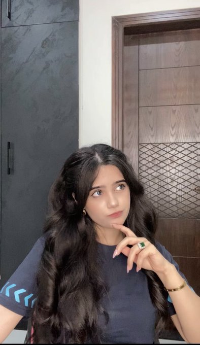 A young woman with long dark wavy hair posing thoughtfully with her hand on her chin, wearing a dark blue t-shirt with blue chevron details on the sleeve and a green gemstone ring. She is photographed indoors in front of a wooden door and dark wardrobe.