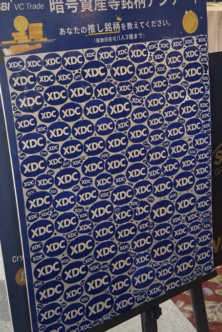 The image shows a promotional board covered in repeating "XDC" logos with Japanese text at the top, tied to the #XDC hashtag representing the XDC Network cryptocurrency.