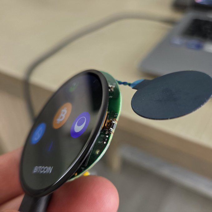 A hand holds a small round electronic device displaying a Bitcoin interface, with an exposed circuit board and a black oval component connected by a wire. The image showcases hardware development progress, tying into the post's excitement about building physical tech for the Sui community through the "Suiball" project.