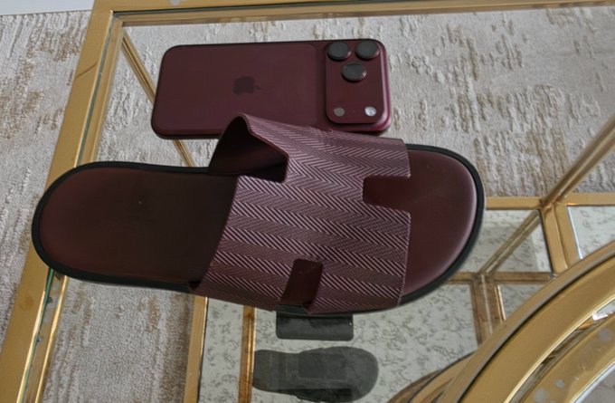 A burgundy Hermès "H" slide sandal rests on a glass table next to a matching burgundy iPhone (likely the "iPhone 18 Pro Max" referenced), highlighting the dramatic price equivalence between the luxury leather slipper and the latest flagship smartphone. The image visually compares the two similarly colored high-end items, reinforcing the question of which expensive purchase one would choose.