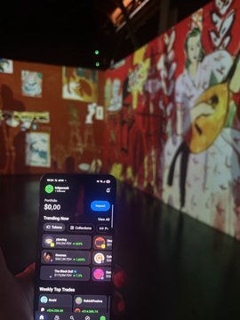 A hand holds a smartphone displaying a crypto app with a balance of $0.00 and 'Trending Now' NFTs, set against a backdrop of colorful digital artwork projected on walls in a dark room.