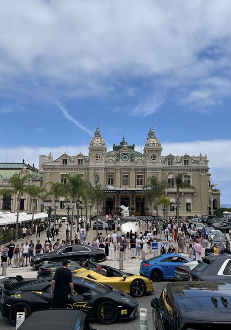 A lineup of luxury supercars, including Ferraris and Lamborghinis, is parked in front of the iconic Monte Carlo Casino in Monaco. The scene perfectly captures the opulent lifestyle referenced in the post, showcasing the lavish travel destinations like Monaco where "crypto kids" are brought instead of a traditional workplace.