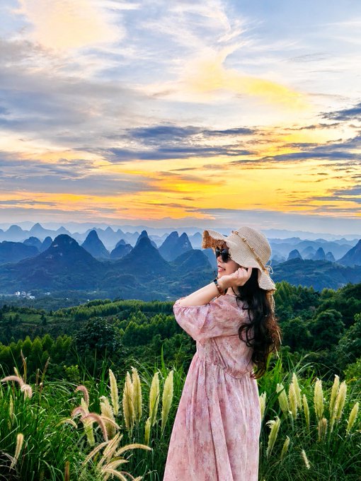 A woman in a pink floral dress and straw hat takes a photo with her phone while standing in a field of tall grass, overlooking a landscape of karst mountains during a vibrant sunset.
