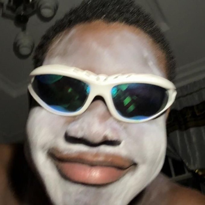 Close-up selfie of a person wearing white-rimmed sunglasses, with their face covered in a white cream or clay mask.