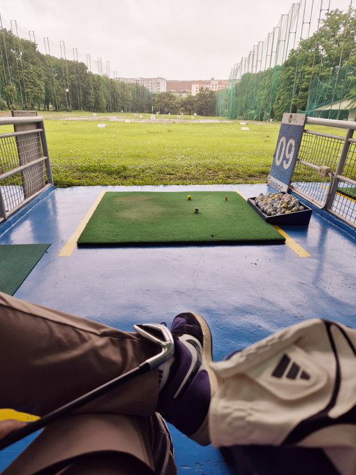 A first-person view from a covered driving range bay showing a person's legs and golf shoes resting while looking out toward a green practice mat marked '09' and a grassy field beyond.
