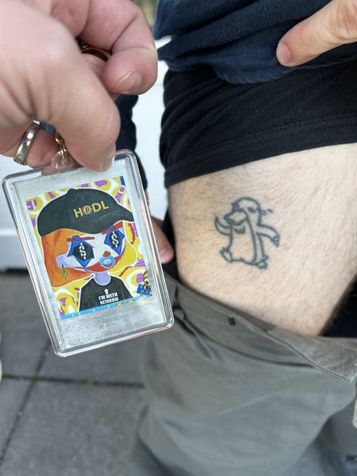 A person holds a 'I'm Built Different' NFT card next to their torso, which displays a matching tattoo of the same character on their skin.
