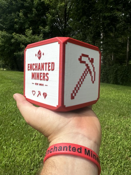 A hand holds a red and white 3D-printed cube featuring 'ENCHANTED MINERS EST. 2024' alongside a pixelated pickaxe design, with the person wearing a matching 'Enchanted Miners' wristband.