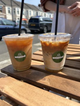 Two iced coffee drinks in clear plastic cups with 'Unsound' logos sit on a wooden picnic table outdoors.
