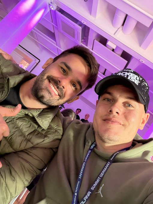 Two smiling young men take a selfie at an indoor event bathed in purple lighting; the one on the right wears a cap and a lanyard reading 'TECH WEEK SUMMIT GERMANY'.