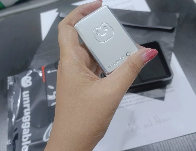 A close-up of a hand holding a silver square Unruggable wallet device with an embossed logo, displayed above a matching branded box and paperwork.