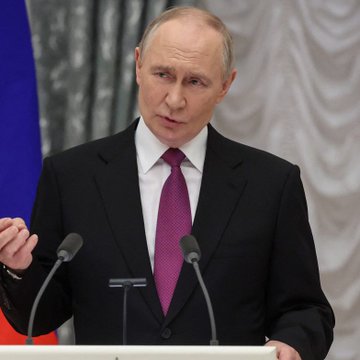 A man with short grey hair, wearing a black suit and magenta tie, speaks at a podium with two microphones against a backdrop of cream curtains and a partial flag. The image likely depicts a Russian official delivering an address related to the post's news about Russia's anticipated growth of up to 10 million new crypto users by 2027 as banks expand regulated crypto trading access.