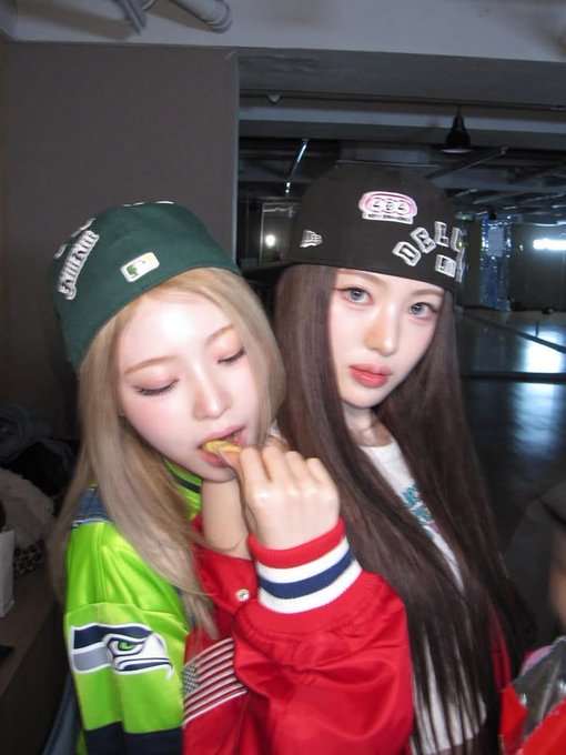 Two young women pose together indoors; one wearing a green baseball cap and a neon green and blue jersey eats a snack while wearing a red and blue striped jacket, and the other wears a black cap with patches and a red jacket.