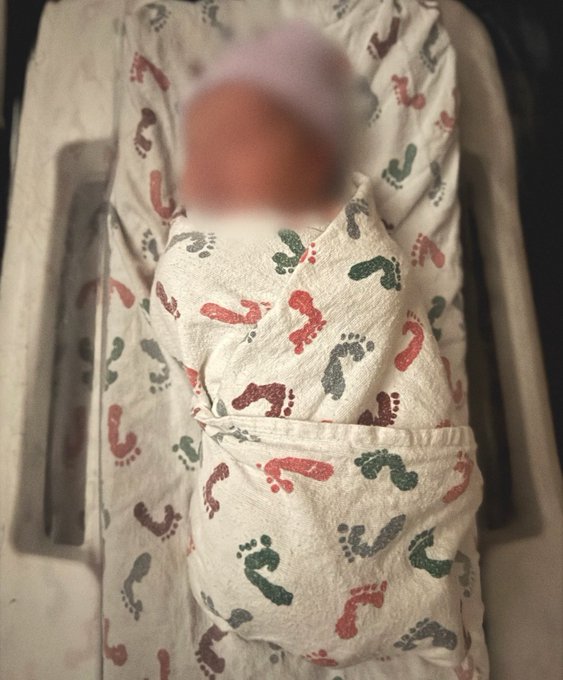 A baby wrapped in a swaddle and hat with a footprint pattern, lying on a matching fabric with its face blurred.