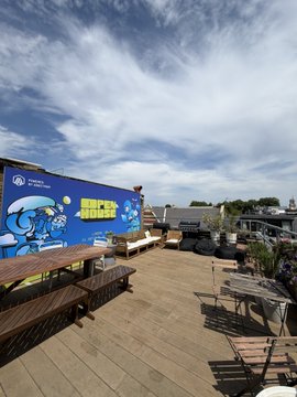 An outdoor rooftop patio or deck area with wooden tables and benches, next to a large blue wall with a mural that includes the text 'FORTITUDE'.